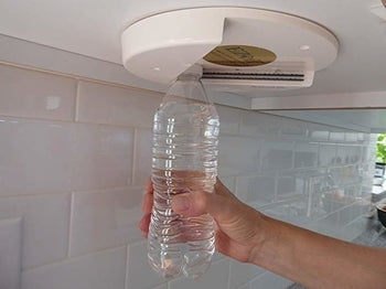 Model's hand putting a plastic water bottle in the corner of the jar opener that's attached to the underside of a cabinet to open the lid