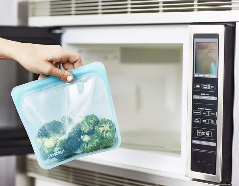 Blue storage bag containing broccoli being placed in microwave 