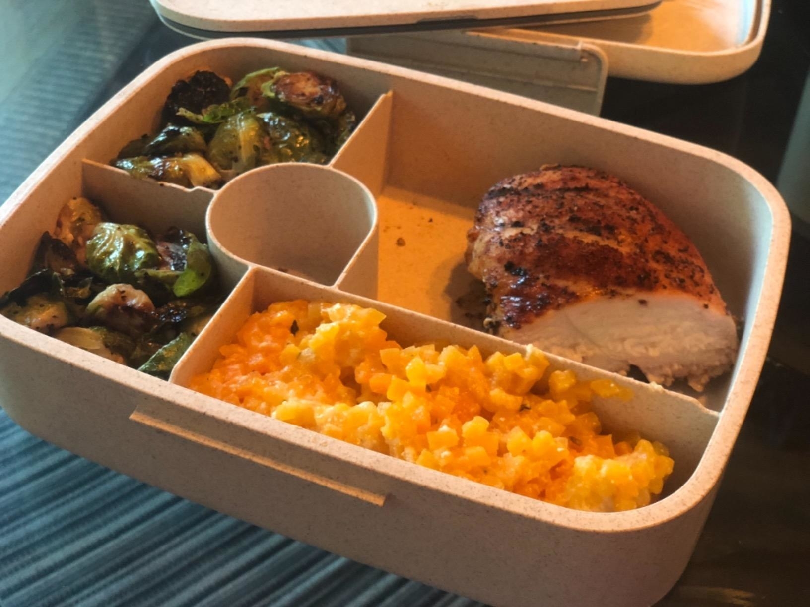 A reviewer&#x27;s photo of the bento box with chicken, risotto, and Brussel sprouts in the different size compartments