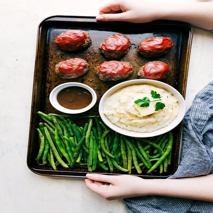 Baked meatloaf with glaze, green beans, mashed potatoes topped with butter and parsley, and gravy on the side, served on a large tray with hands holding it