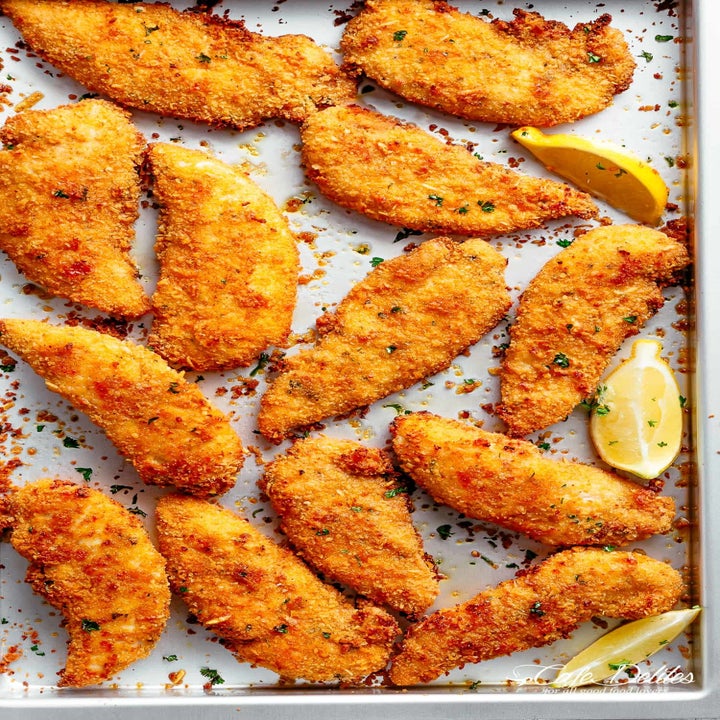 A baking tray filled with golden, crispy breaded chicken tenders garnished with herbs and accompanied by lemon wedges