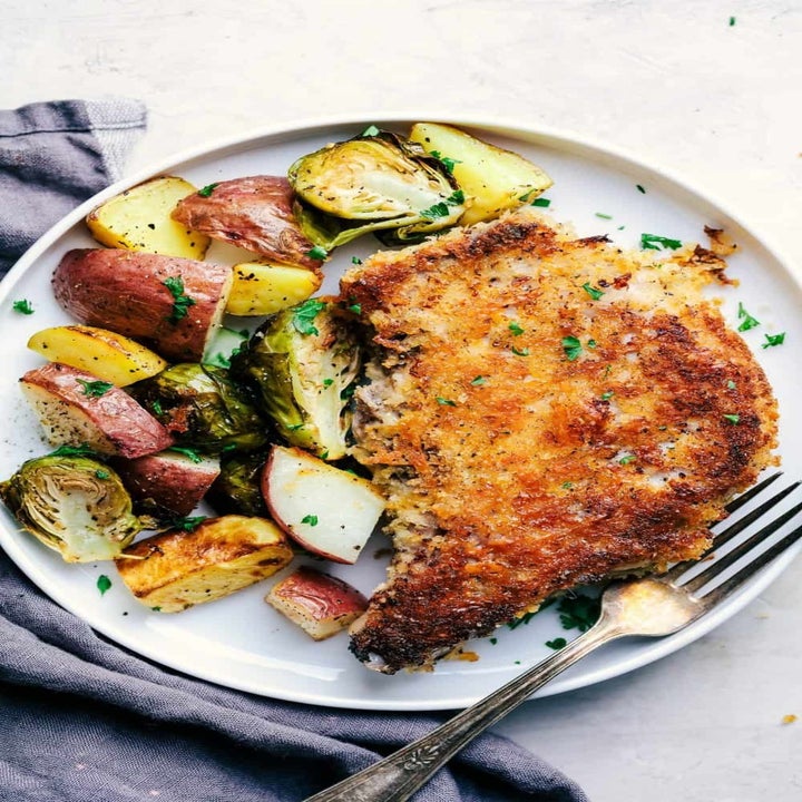 A plate of crispy breaded pork chop served with roasted potatoes and Brussels sprouts, garnished with herbs. A fork rests on the plate