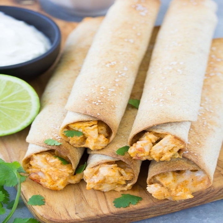 Four chicken taquitos on a wooden board, garnished with fresh cilantro, accompanied by a lime wedge and a bowl of white dip