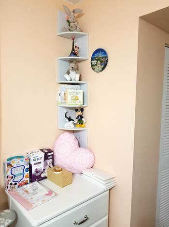 a skinny corner shelf with five tiers in white holding baby items in a nursery