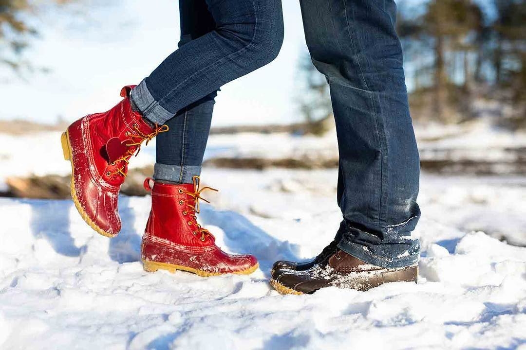 two model&#x27;s feet in the snow, one in a red pair and one a brown pair of the same high-top boot; there&#x27;s rubber around the toes and bottom third of the shoes