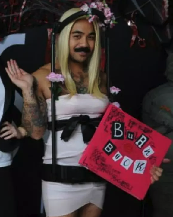 Someone with a mustache dressed as Regina George in her Spring Fling dress while holding the Burn Book