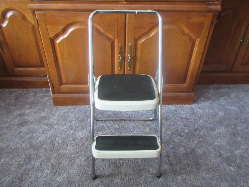 A two step step stool opened and in front of a wooden cabinet 