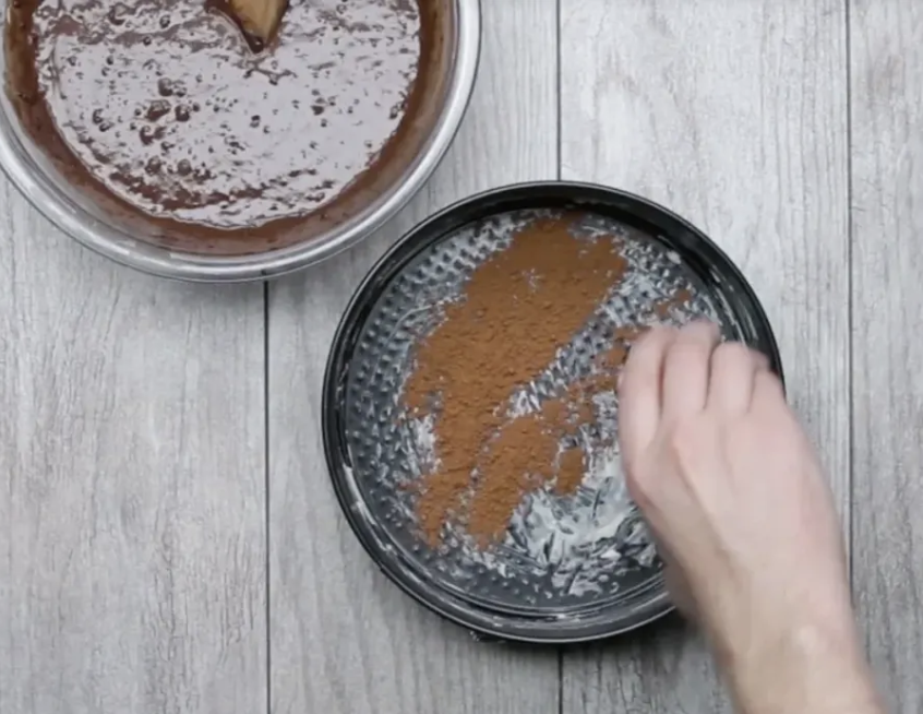 A baker sprinkling a bit of a cocoa powder into a prepped cake pan