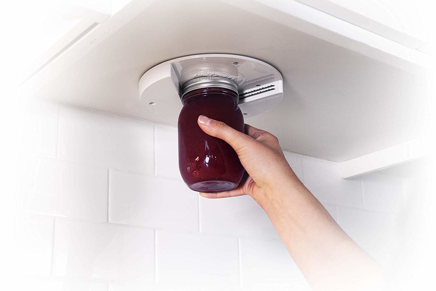 A model using a white round jar opener mounted under a cabinet to pull a lid off a jar 