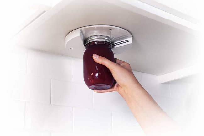 A model using a white round jar opener mounted under a cabinet to pull a lid off a jar 