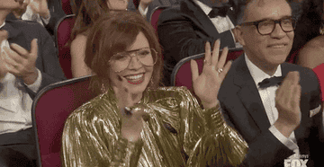 Natasha in a shimmering outfit smiling and clapping awkwardly