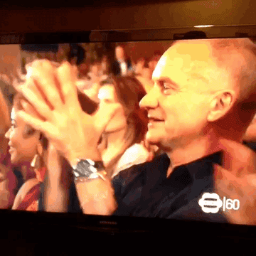 Sting applauding at an event, wearing a dark shirt and watch, with his fingers far apart