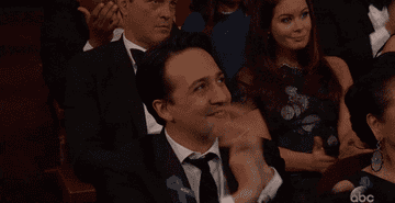 Lin-Manuel in a black suit clapping but with one hand almost missing the other