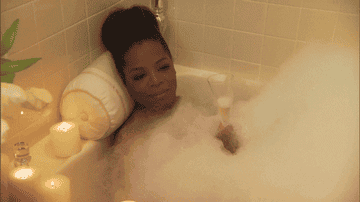 A GIF of Oprah Winfrey sitting in a bathtub holding a glass of wine and covered in bubbles