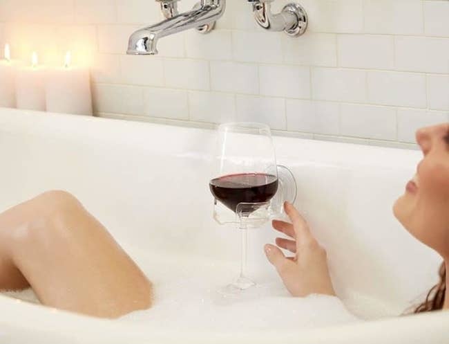 Model in a bubble bath with the clear holder suctioned to the side of the tub with a glass of red wine in it. 