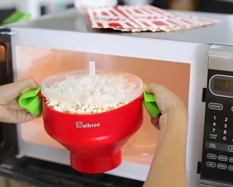 The red bowl with a clear top with popped popcorn in it and a model&#x27;s hands pulling it out of a microwave. 