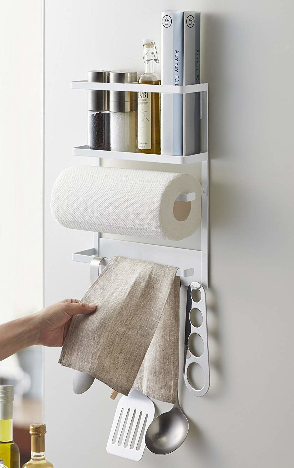 organizer on side of a fridge holding a paper towel roll, salt, pepper, and oil on a top rack, and a towel and cooking utensils hanging from hooks on the bottom