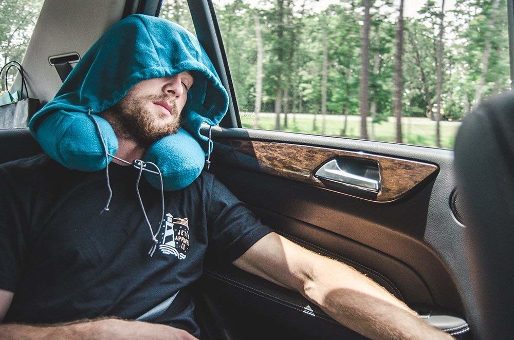 A man riding in the back seat of a car, taking a nap while wearing the hooded travel pillow in peacock green