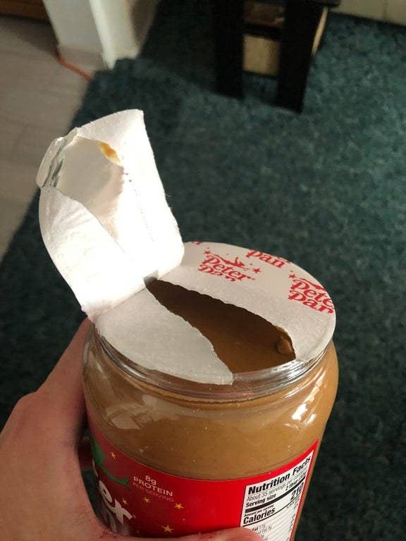 Jar of peanut butter with a foil seal partially removed, held in a hand