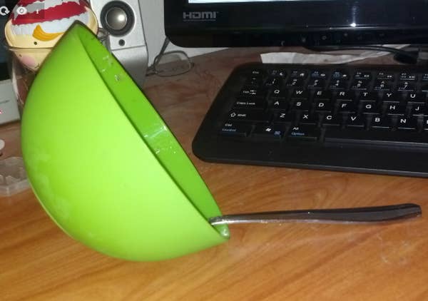 A green bowl with a spoon balanced on a desk in front of a keyboard and a monitor