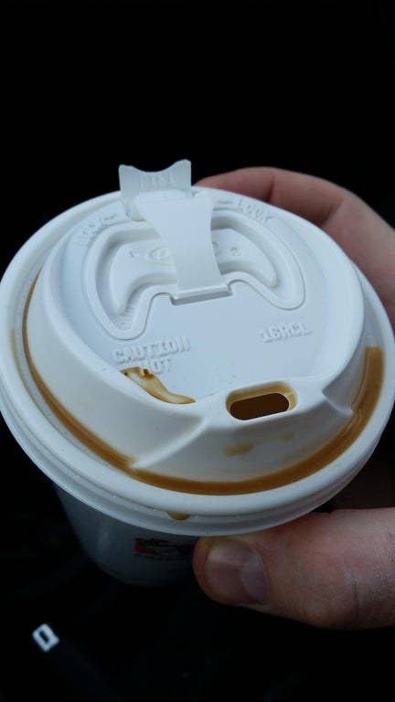 A hand holding a disposable coffee cup with a plastic lid, showing spilled coffee around the rim