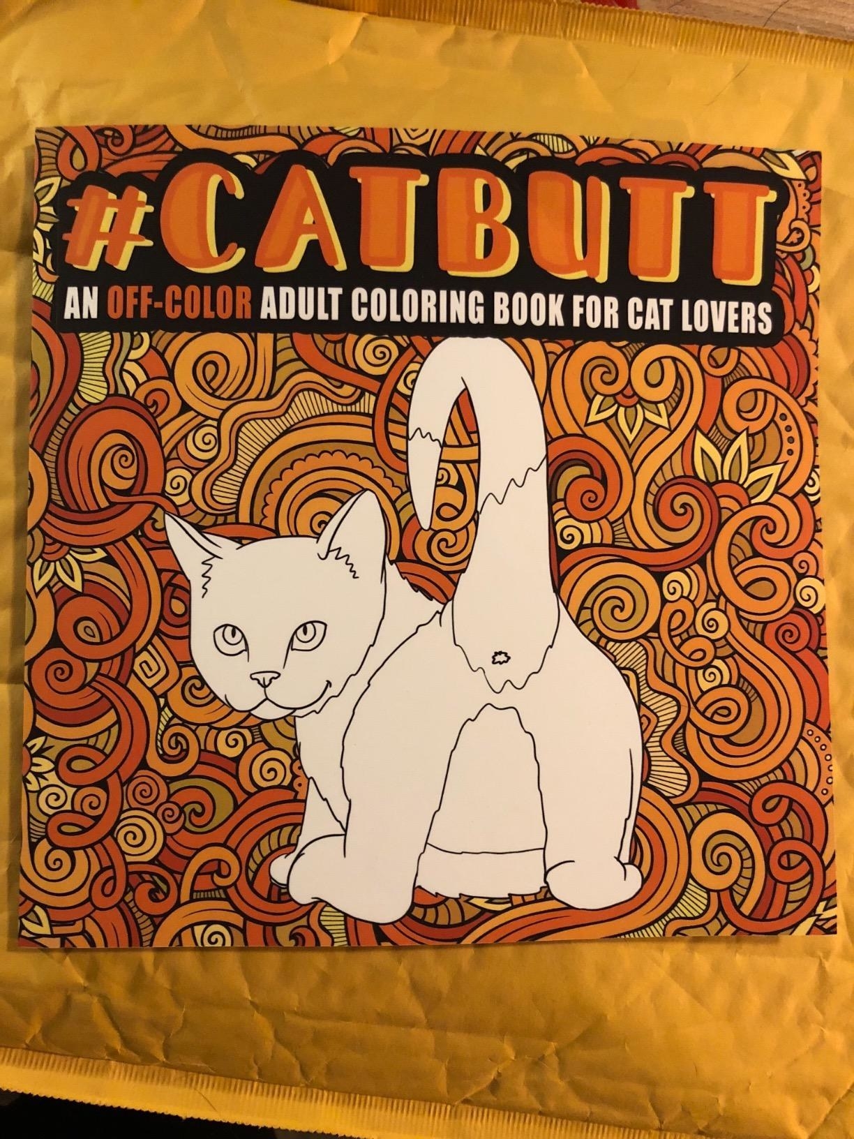A reviewer&#x27;s photo of the coloring book cover, which shows a white ready-to-be-colored cat showing its butt and looking cheekily at the viewer with the title &quot;#Catbutt&quot; in top