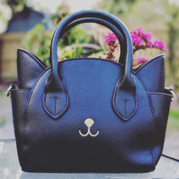 Reviewer pic of the purse in black with ears on either side and pockets below them and a small white nose and mouth on the front with the ends of the straps looking like eyes. 