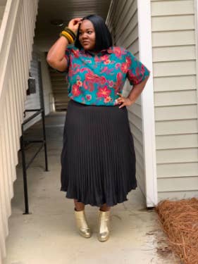 plus size reviewer wearing the black pleated ankle length skirt with a floral top and gold ankle boots
