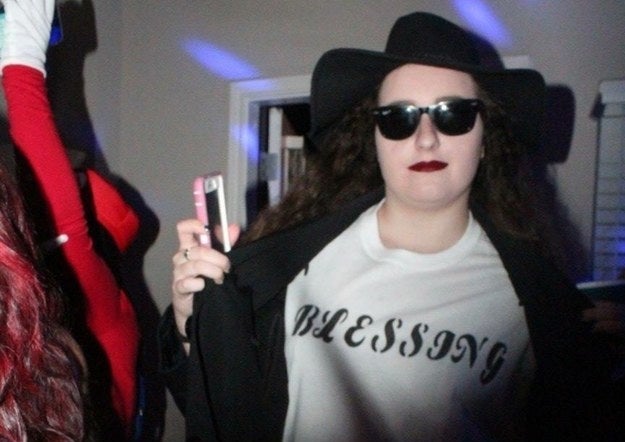 A woman in a shirt that says &quot;blessing&quot; that is covered by a large coat, sunglasses, and a hat