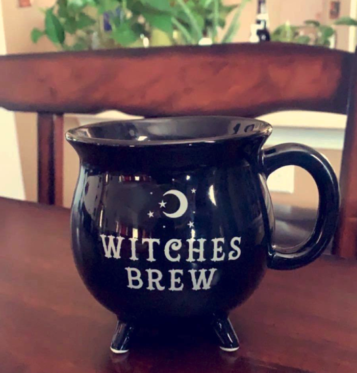 a black mug shaped like a cauldron with four legs on the bottom and a handle. It says, &quot;witches brew.&quot; 