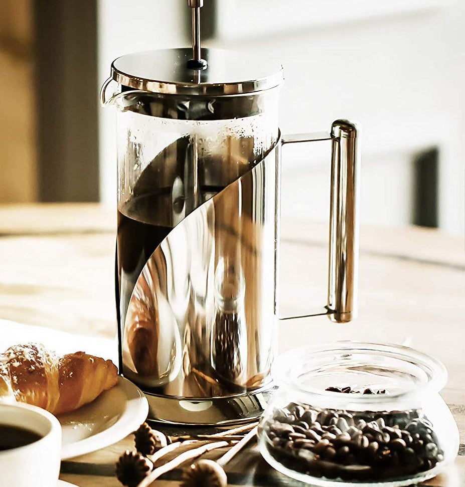 A metal French press on a table next to a glass jar full of coffee beans and a plate with a croissant on it