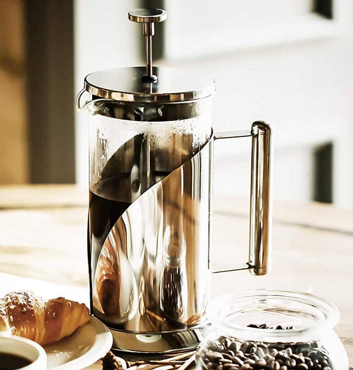 A metal French press on a table next to a glass jar full of coffee beans and a plate with a croissant on it