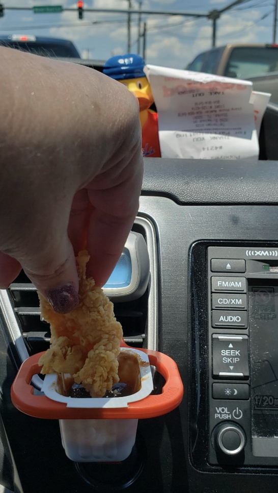 reviewer pic of person dipping a chicken tender into tthe little clip attached to a car vent with the sauce container inside