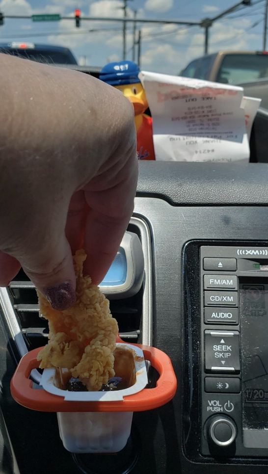 reviewer pic of person dipping a chicken tender into tthe little clip attached to a car vent with the sauce container inside