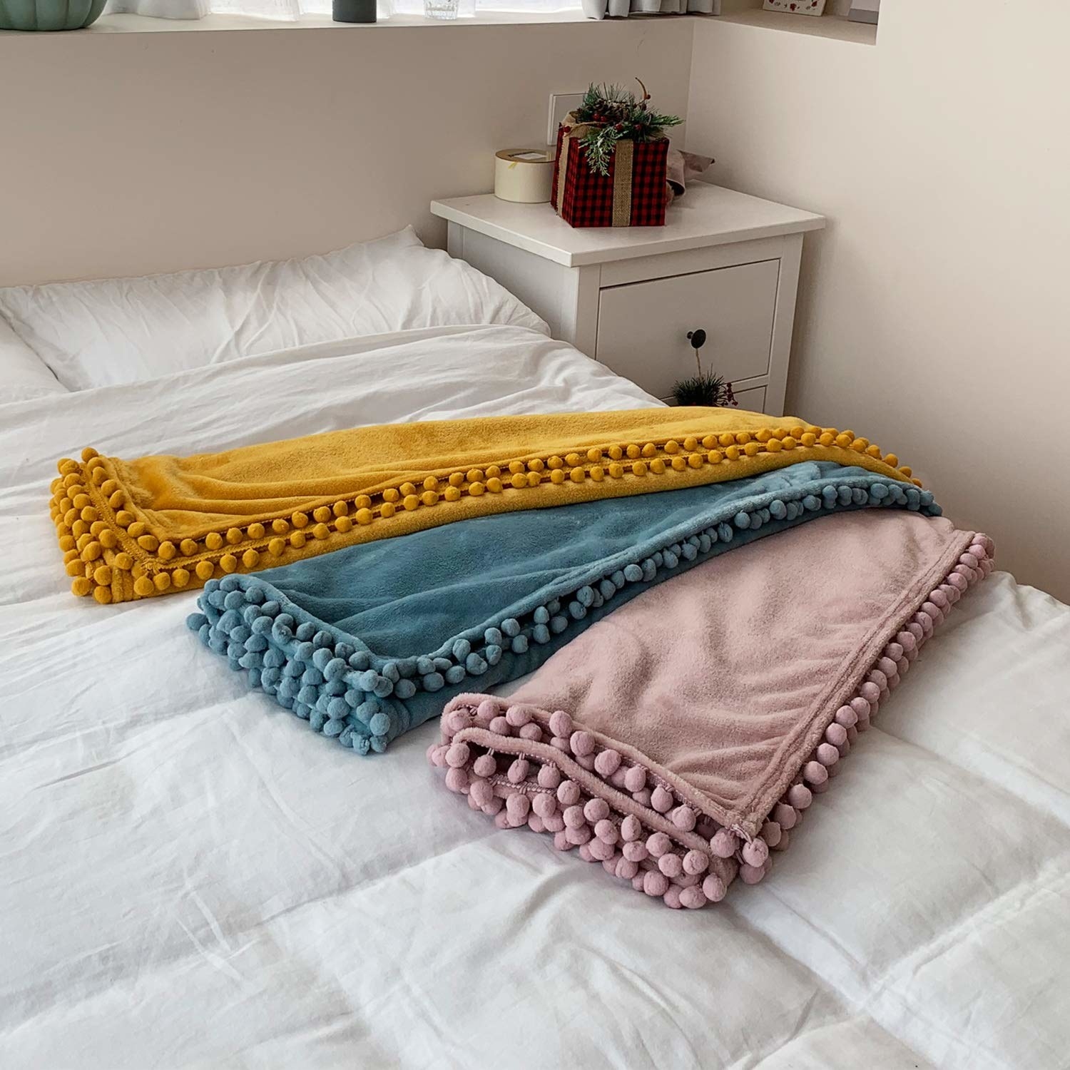 Three blankets in different colors with pom pom edges tossed across white bedding 