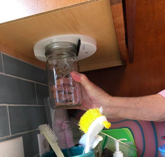 A reviewer placing a mason jar lid into the disk-shaped opener, which is mounted under their cabinet