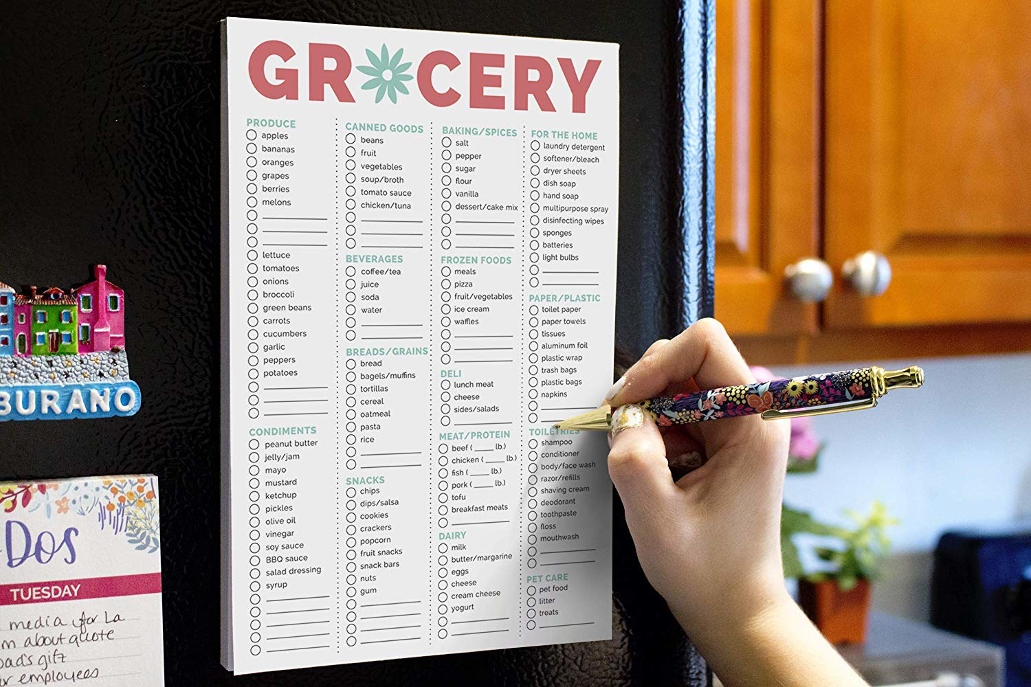 The grocery pad stuck to the side of the fridge and a hand check-marking common items needed