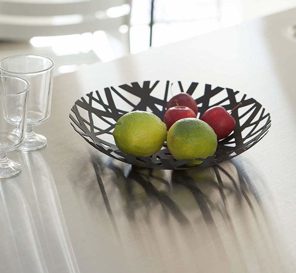 Black bowl made of crisscrossing stripes with openings and assorted limes and other fruit inside