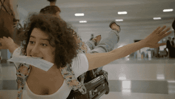 gif of Ilana Glazer in the TV show &quot;Broad City&quot; on top of a suitcase with her arms stretched behind her and a ticket in her mouth
