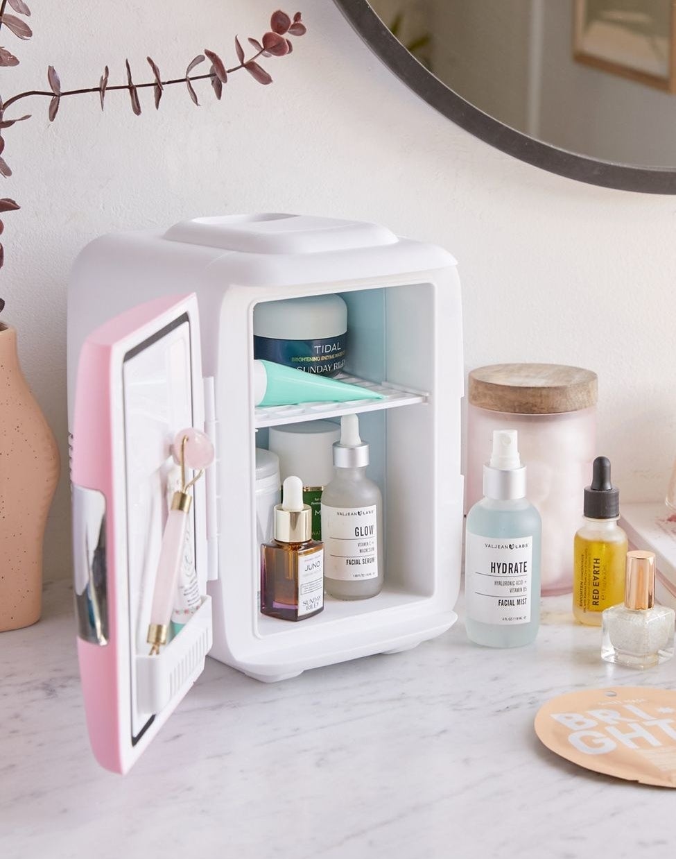 The small fridge on a counter with a pink door open, showing how it has a shelf on the inside of the door, and one in the fridge that&#x27;s filled with assorted beauty products
