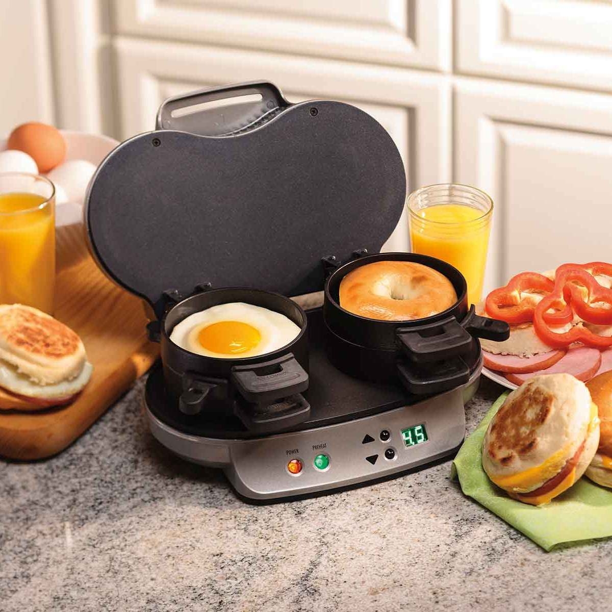 Dual breakfast sandwich maker in silver with egg in the left side, bagel in the right, and other assorted ingredients around it