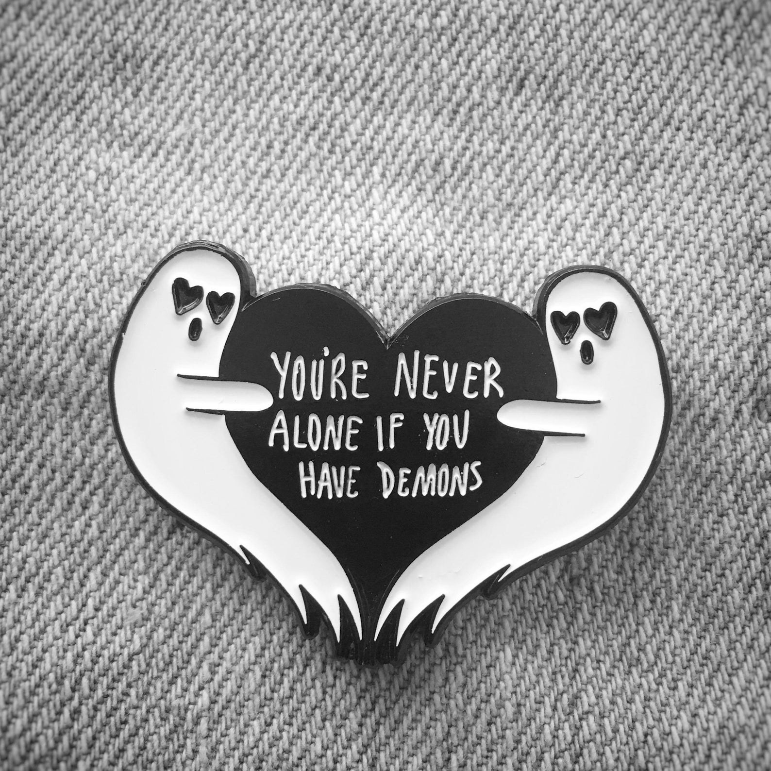 the pin with two ghosts and a black heart in between that says you&#x27;re never alone if you have demons 