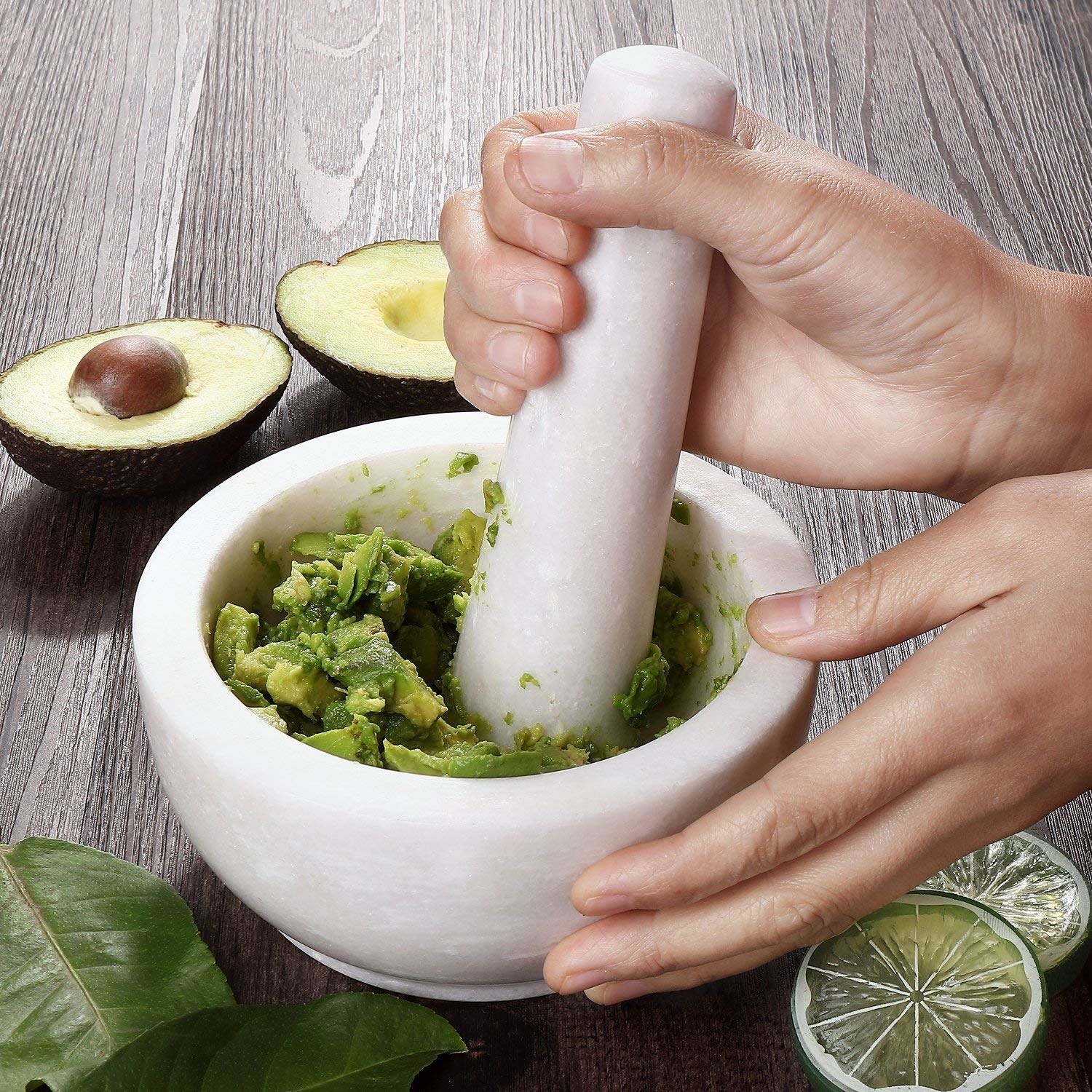 Round white bowl with a hand smashing some avocado with the stick-shaped pestle also in white