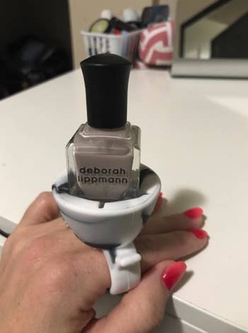 reviewer's hand with the nail polish holder that fits over their hand like a ring. It's holding a bottle of gray nail polish.