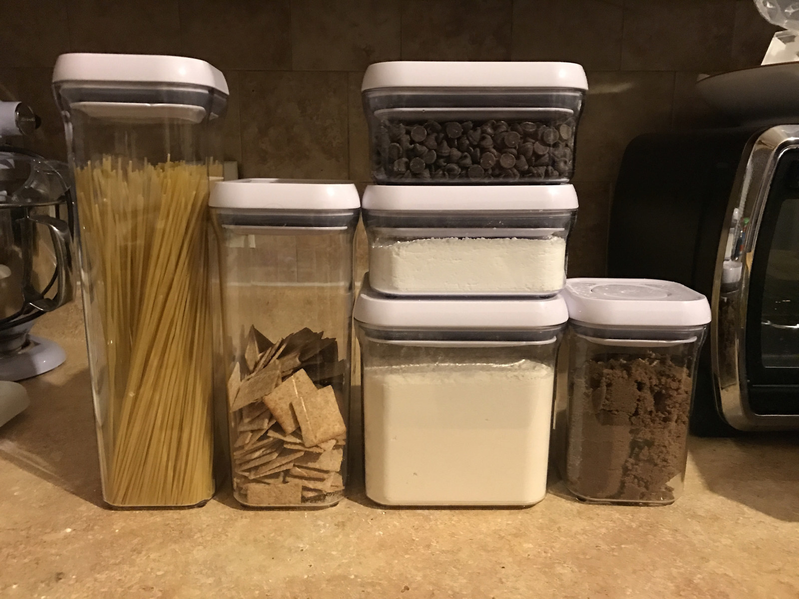 Six OXO Pop Containers of various sizes filled with a mix of dry ingredients from pasta to chocolate chips