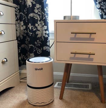 The air purifier in white sitting next to a nightstand