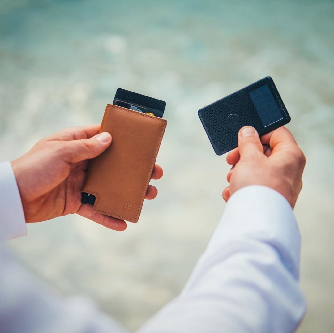 The rectangle-shaped wallet in light brown leather in a model&#x27;s hand with cards coming out of the top and the card-shaped tracker in the other hand