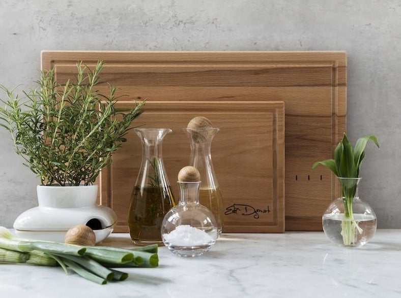 Three glass carafes with round oak stoppers on a countertop next to various leafy plants and in front of two standing wooden cutting boards 