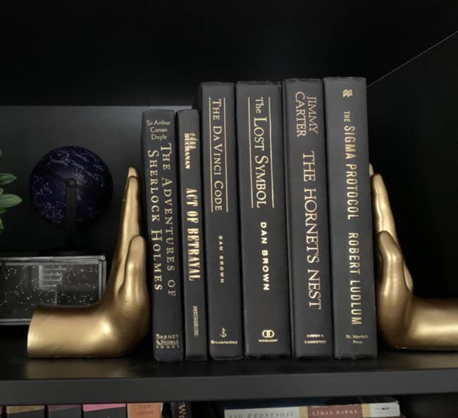 six black books with gold writing on a bookshelf with two hands holding them up