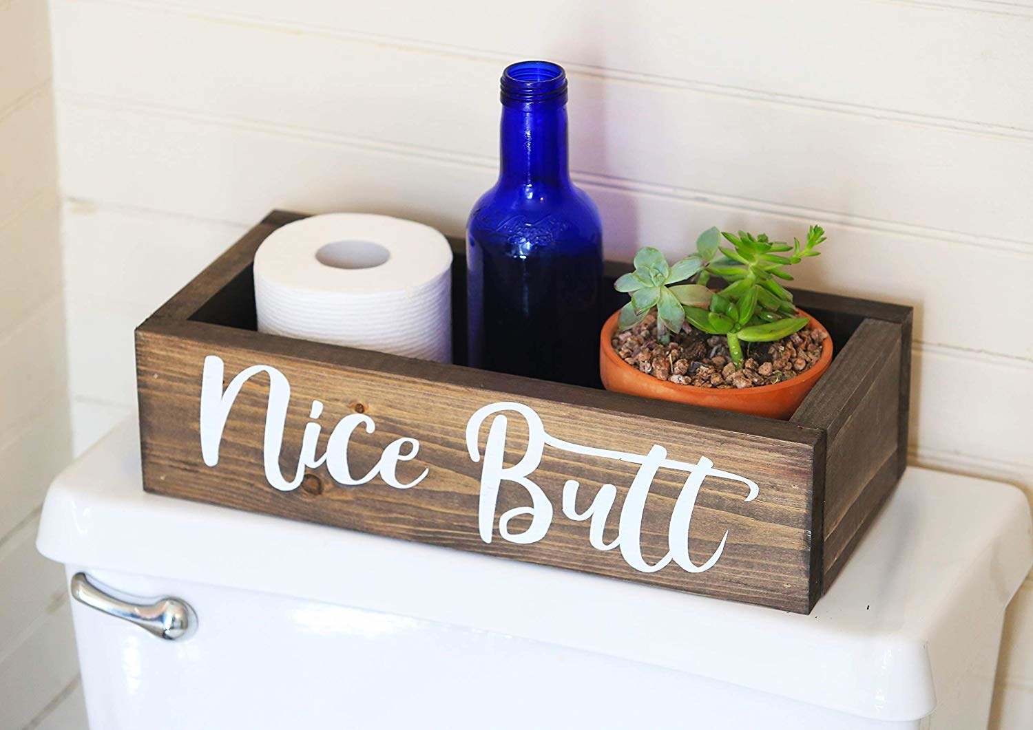 Rectangular wood box on toilet tank holding roll of toilet paper, plant, and bottle with the words &quot;nice butt&quot; painted on the front 